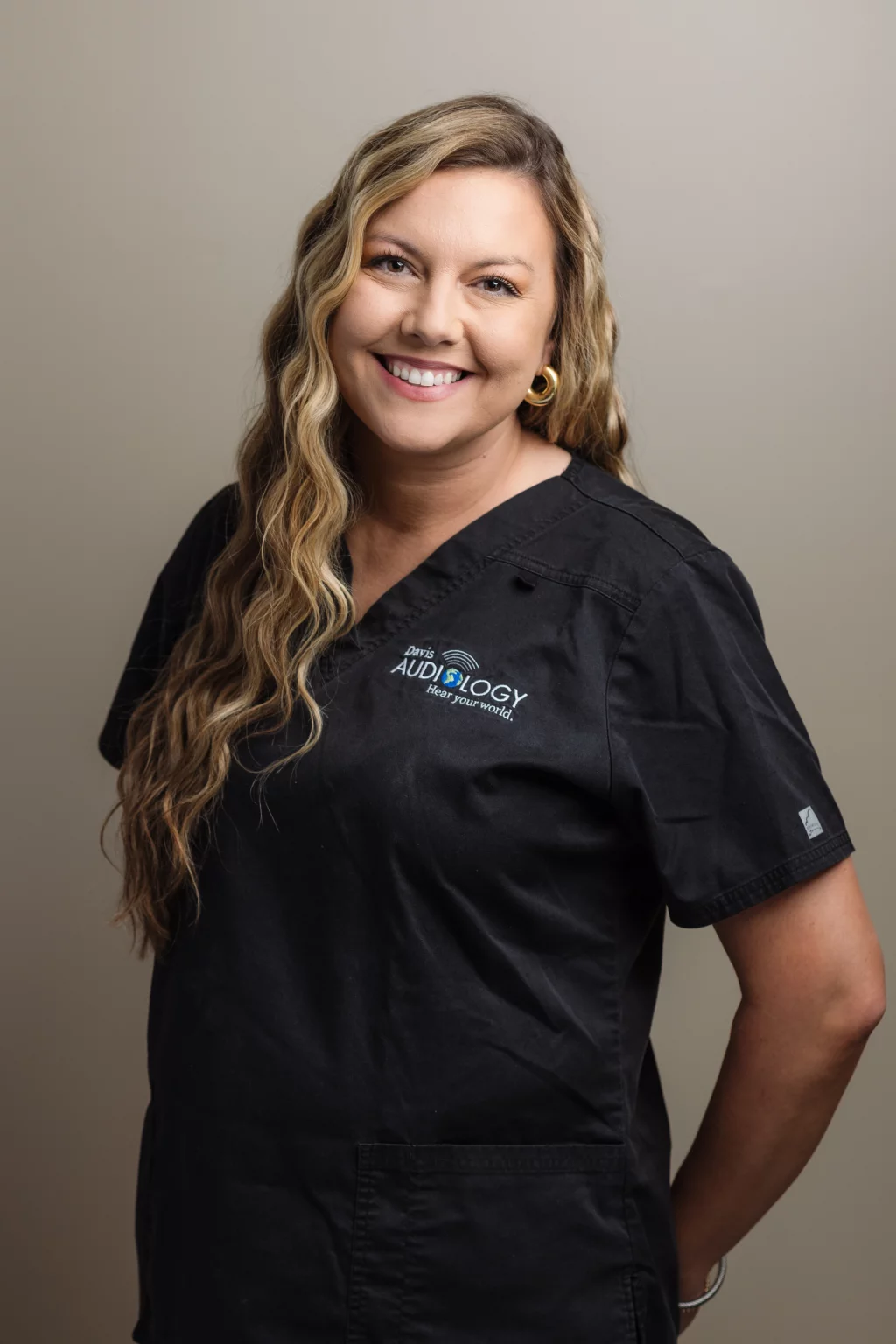 Headshots and branding for Davis Audiology in Taylors SC.: Smiling woman with long wavy blonde hair wearing black medical scrubs embroidered with &ldquo;Davis Audiology,&rdquo; posing against a neutral background.