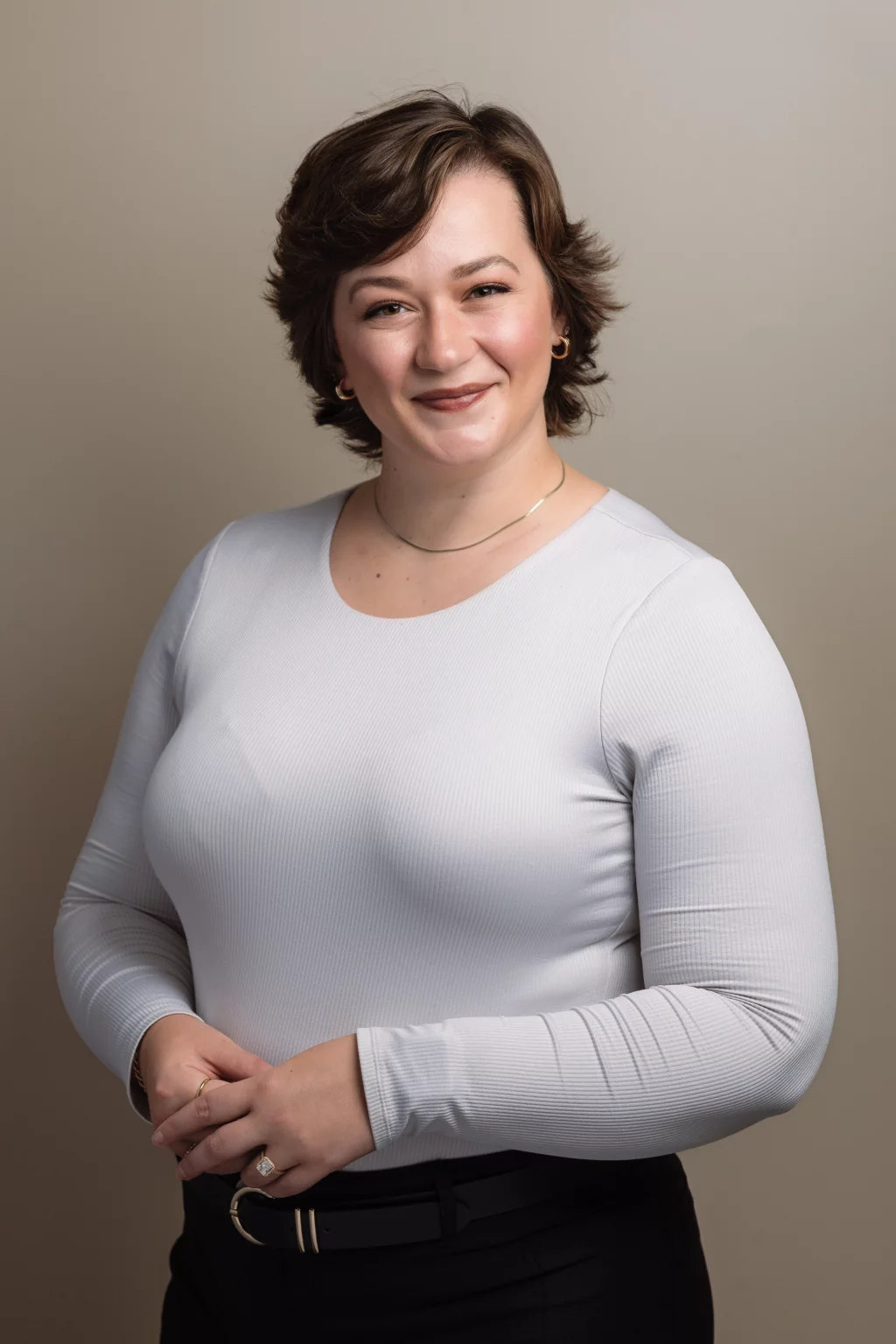 Headshots and branding for Davis Audiology in Taylors SC.: Portrait of a smiling woman with short brown hair, wearing a white long-sleeved top, black pants, gold jewelry, and a belt, standing against a beige background.