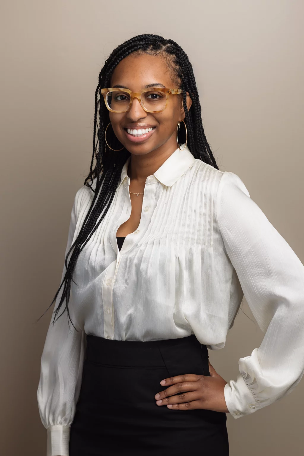 Headshots and branding for Davis Audiology in Taylors SC.: Portrait of a smiling woman with long braids and translucent glasses, wearing a white blouse and black skirt against a beige background.