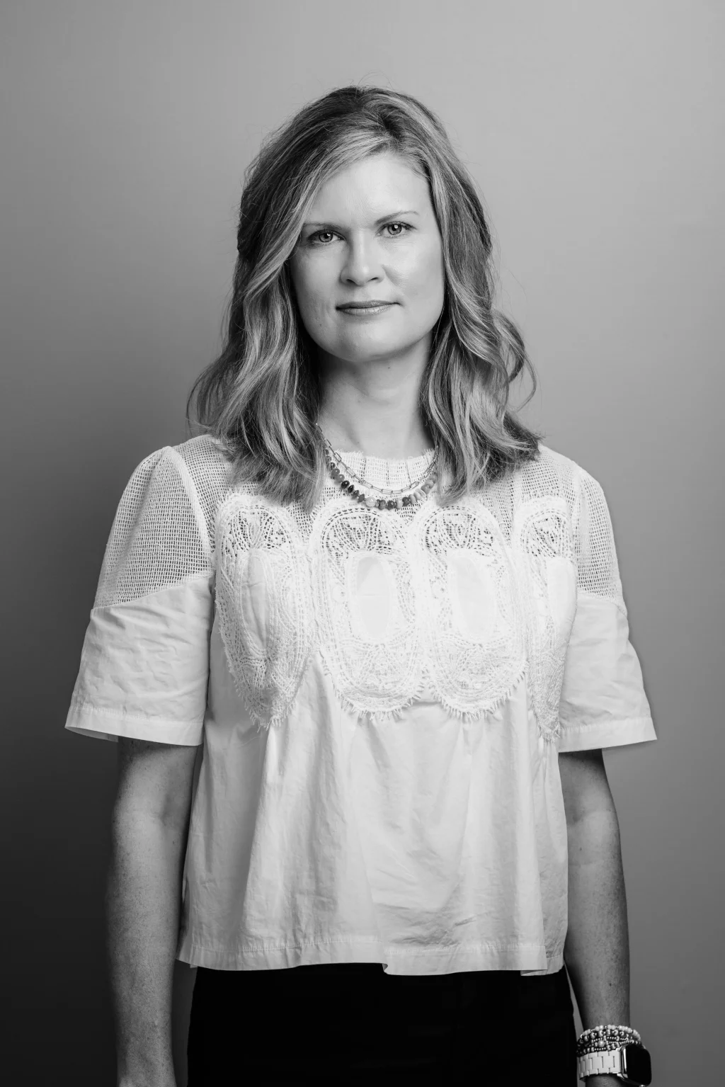 Headshots and branding for Davis Audiology in Taylors SC.: Black-and-white portrait of a woman with shoulder-length wavy hair, wearing a white lace-detailed blouse, layered necklaces, and a smartwatch, against a plain background.