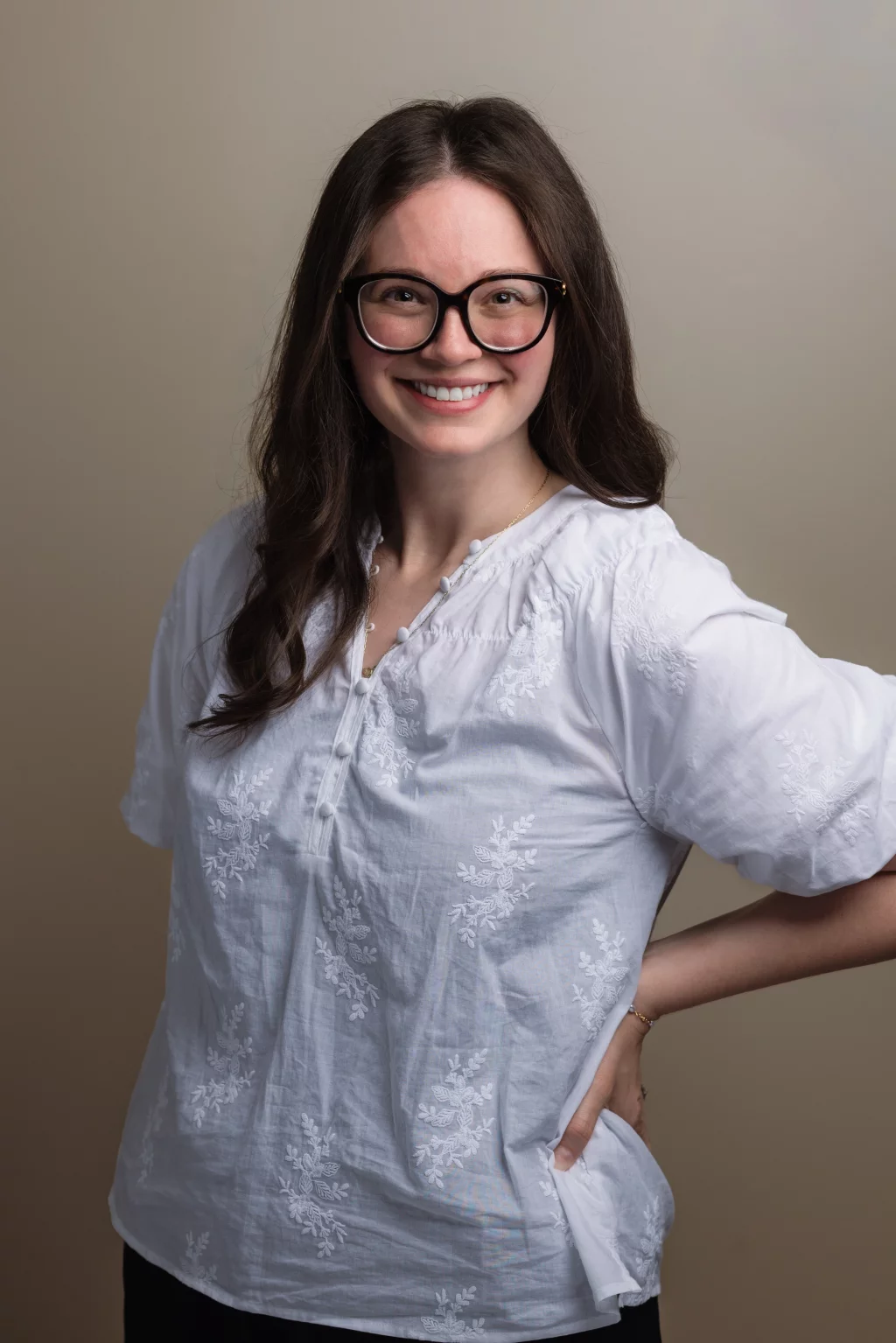 Headshots and branding for Davis Audiology in Taylors SC.: Smiling woman with long dark hair and black glasses wearing a white embroidered blouse, posing with one hand on her hip against a plain brown background.