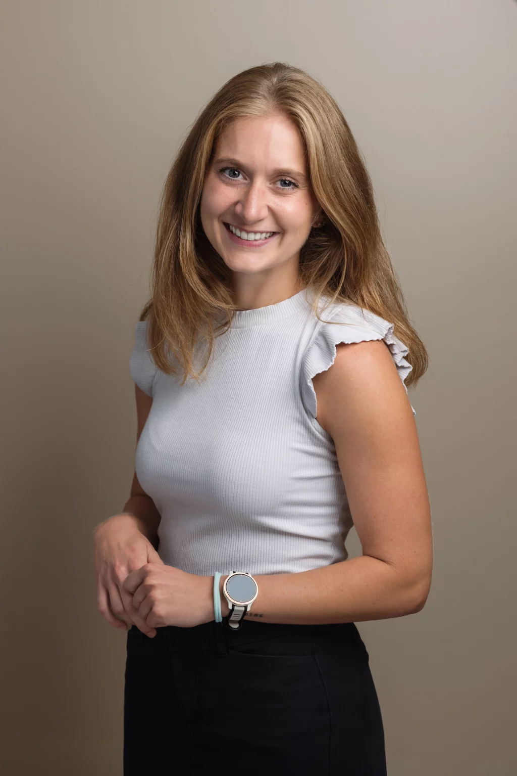 Headshots and branding for Davis Audiology in Taylors SC.: Portrait of a smiling woman with long light-brown hair wearing a white ribbed top, black pants, and a smartwatch, standing against a neutral background.