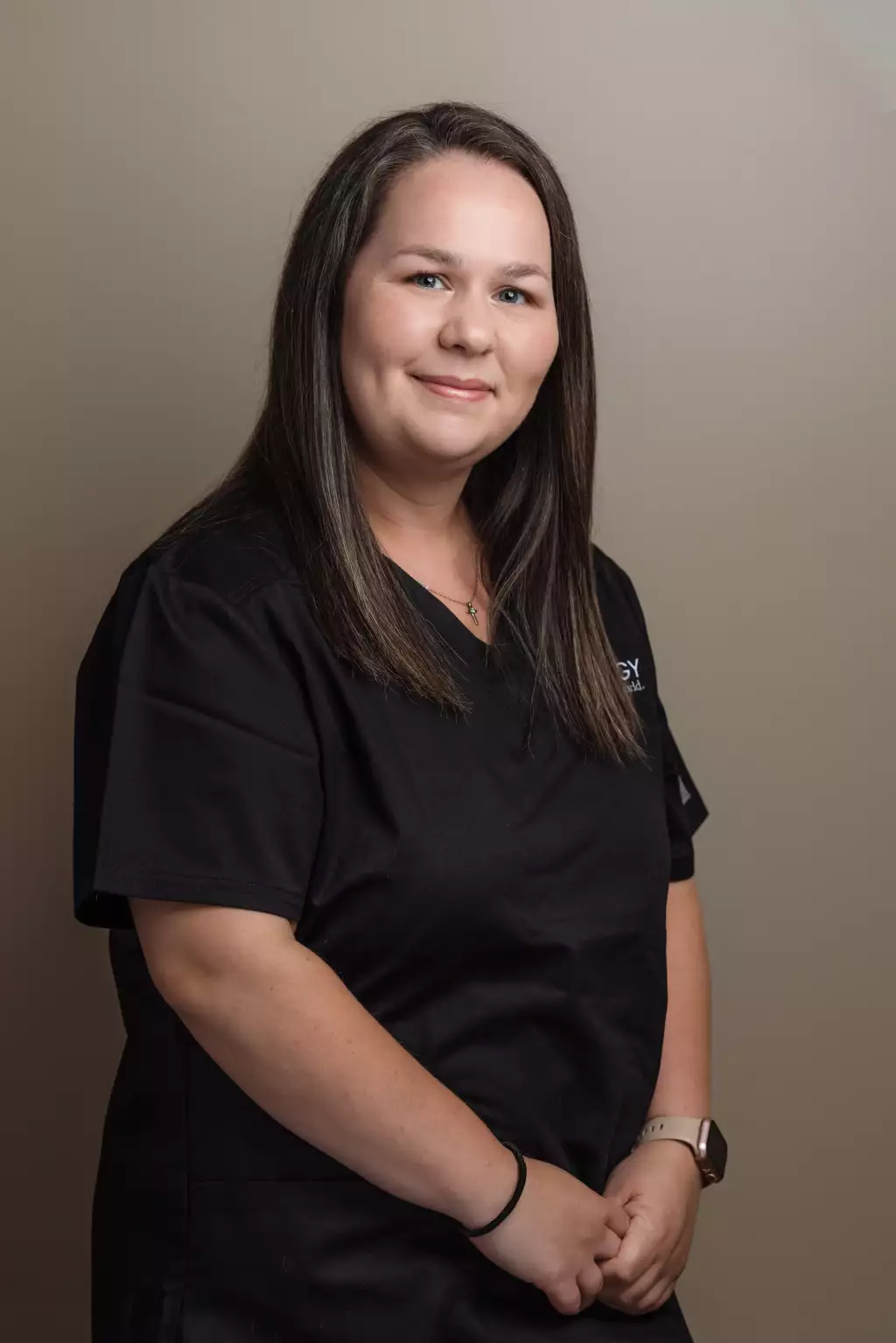 : Portrait of a smiling woman with long dark hair wearing black medical scrubs, a cross necklace, and a smartwatch, standing against a neutral beige background.