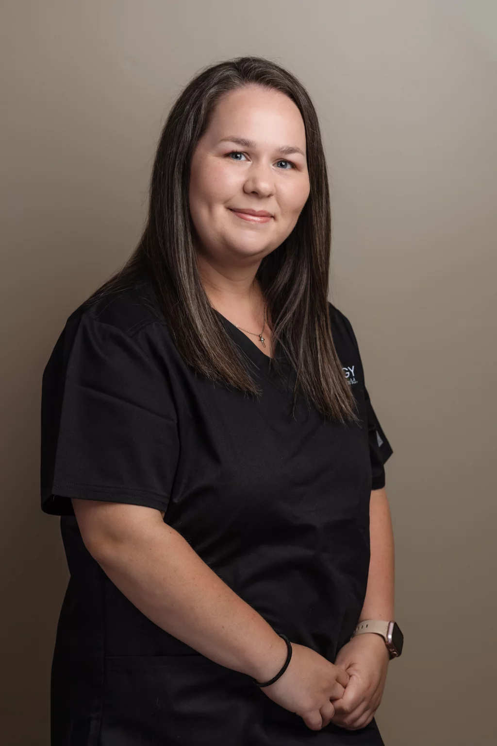Headshots and branding for Davis Audiology in Taylors SC.: Portrait of a smiling woman with long dark hair wearing black medical scrubs, a cross necklace, and a smartwatch, standing against a neutral beige background.