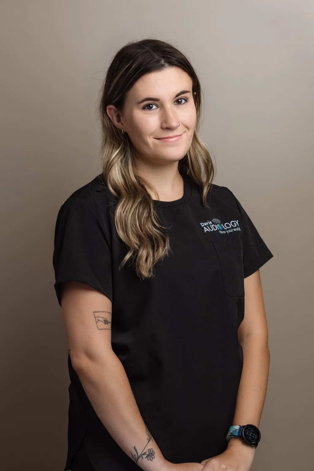 Headshots and branding for Davis Audiology in Taylors SC.: Portrait of a smiling woman with long wavy brown hair wearing black Davis Audiology scrubs, a smartwatch, and visible tattoos, against a neutral background.
