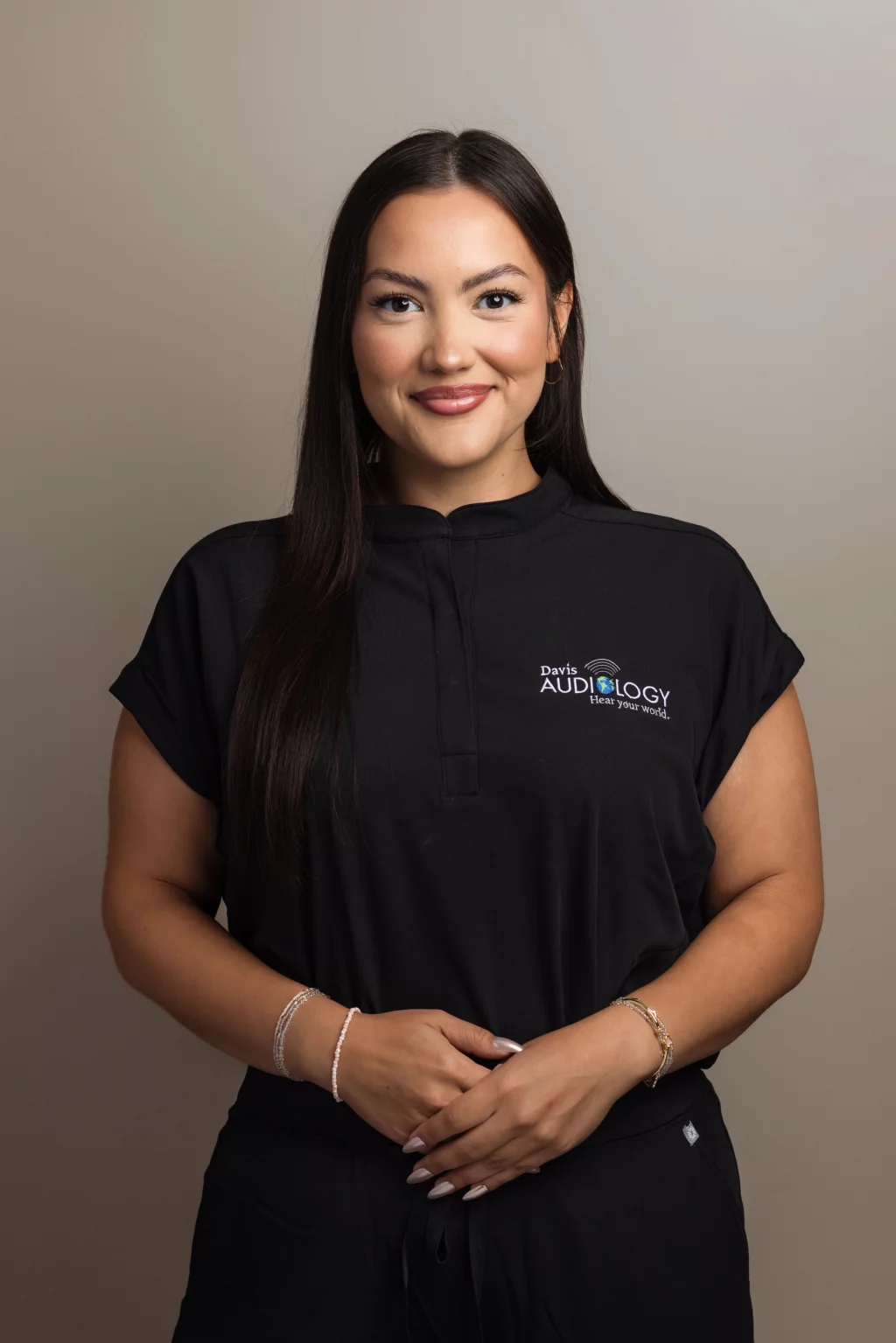 Headshots and branding for Davis Audiology in Taylors SC.: Portrait of a smiling woman with long dark hair wearing a black Davis Audiology uniform, standing with her hands clasped against a neutral background.