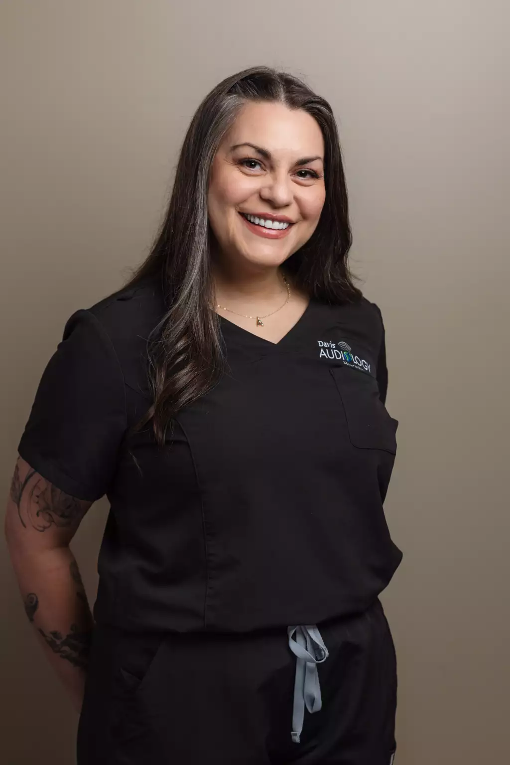 : Smiling woman with long dark hair and visible arm tattoos wearing black medical scrubs with a &ldquo;Davis Audiology&rdquo; logo, posed against a plain beige background.