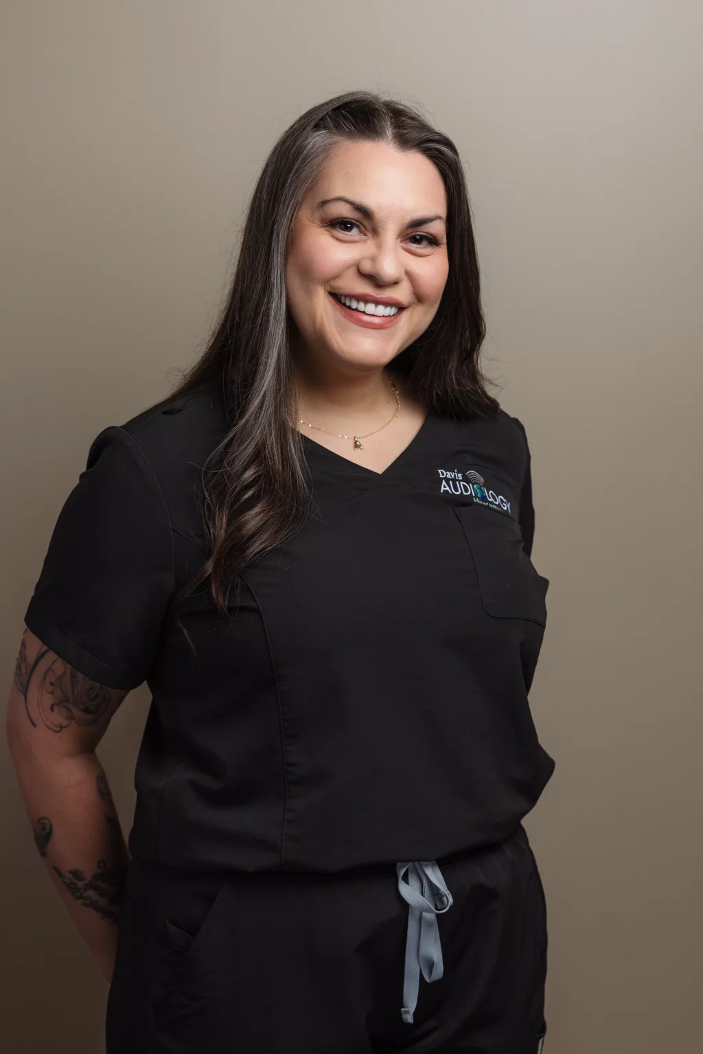 Headshots and branding for Davis Audiology in Taylors SC.: Smiling woman with long dark hair and visible arm tattoos wearing black medical scrubs with a &ldquo;Davis Audiology&rdquo; logo, posed against a plain beige background.