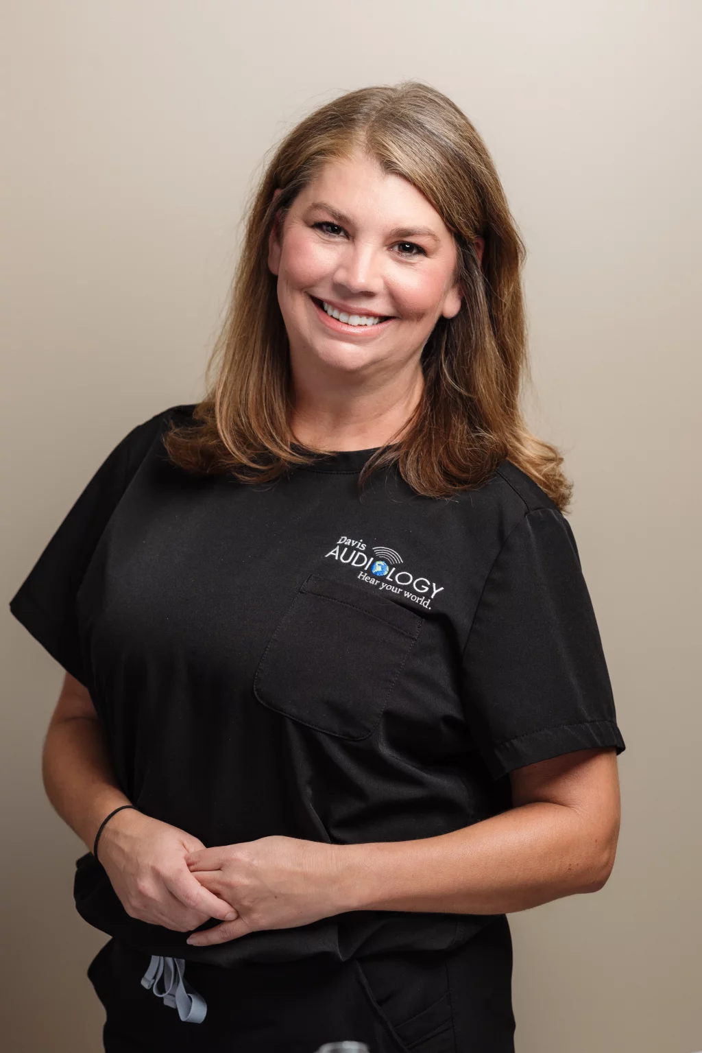 Headshots and branding for Davis Audiology in Taylors SC.: Smiling woman with shoulder-length brown hair wearing black medical scrubs embroidered with &ldquo;Davis Audiology,&rdquo; standing against a plain beige background.