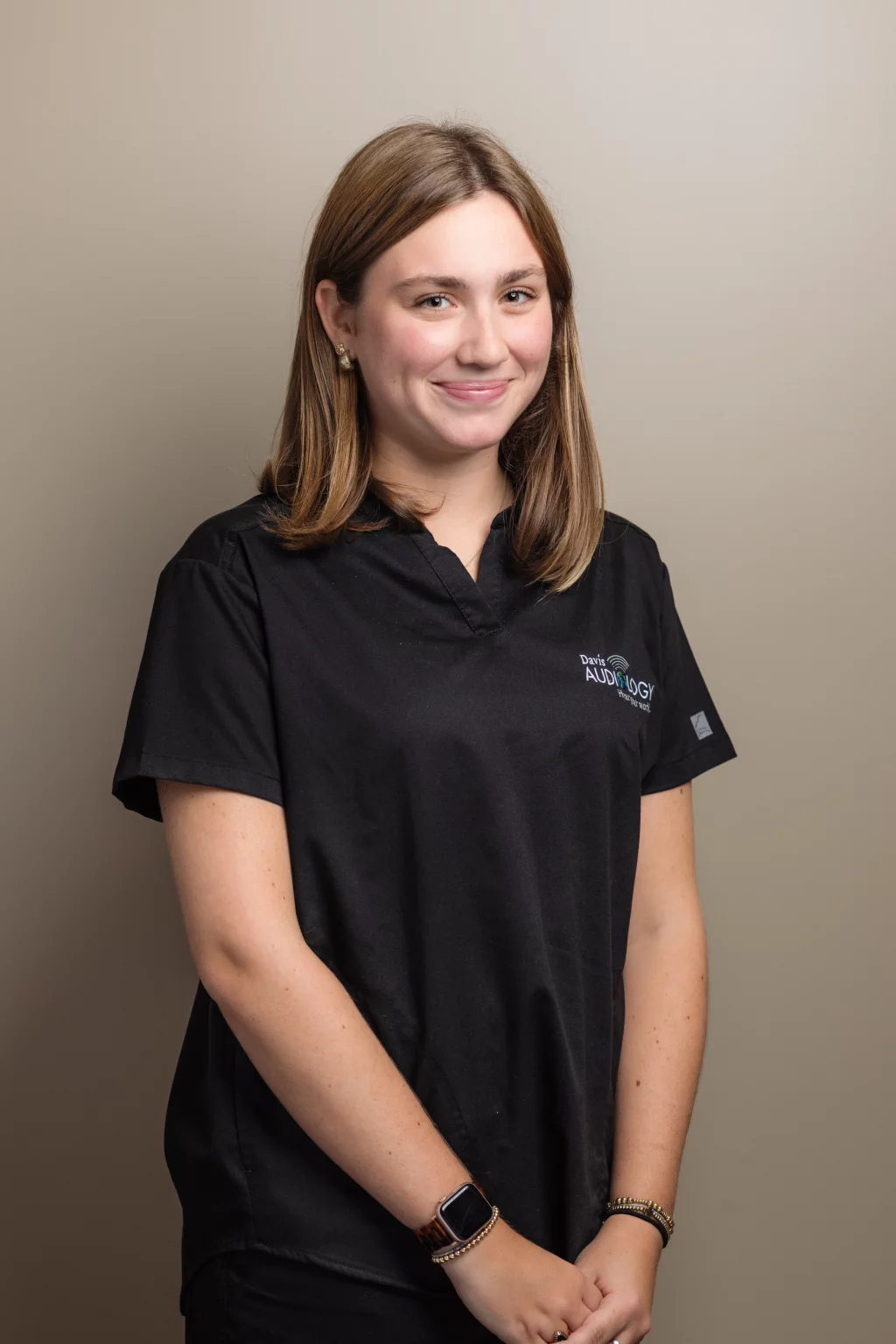 Headshots and branding for Davis Audiology in Taylors SC.: Portrait of a smiling woman with shoulder-length brown hair wearing black medical scrubs, standing against a neutral background.