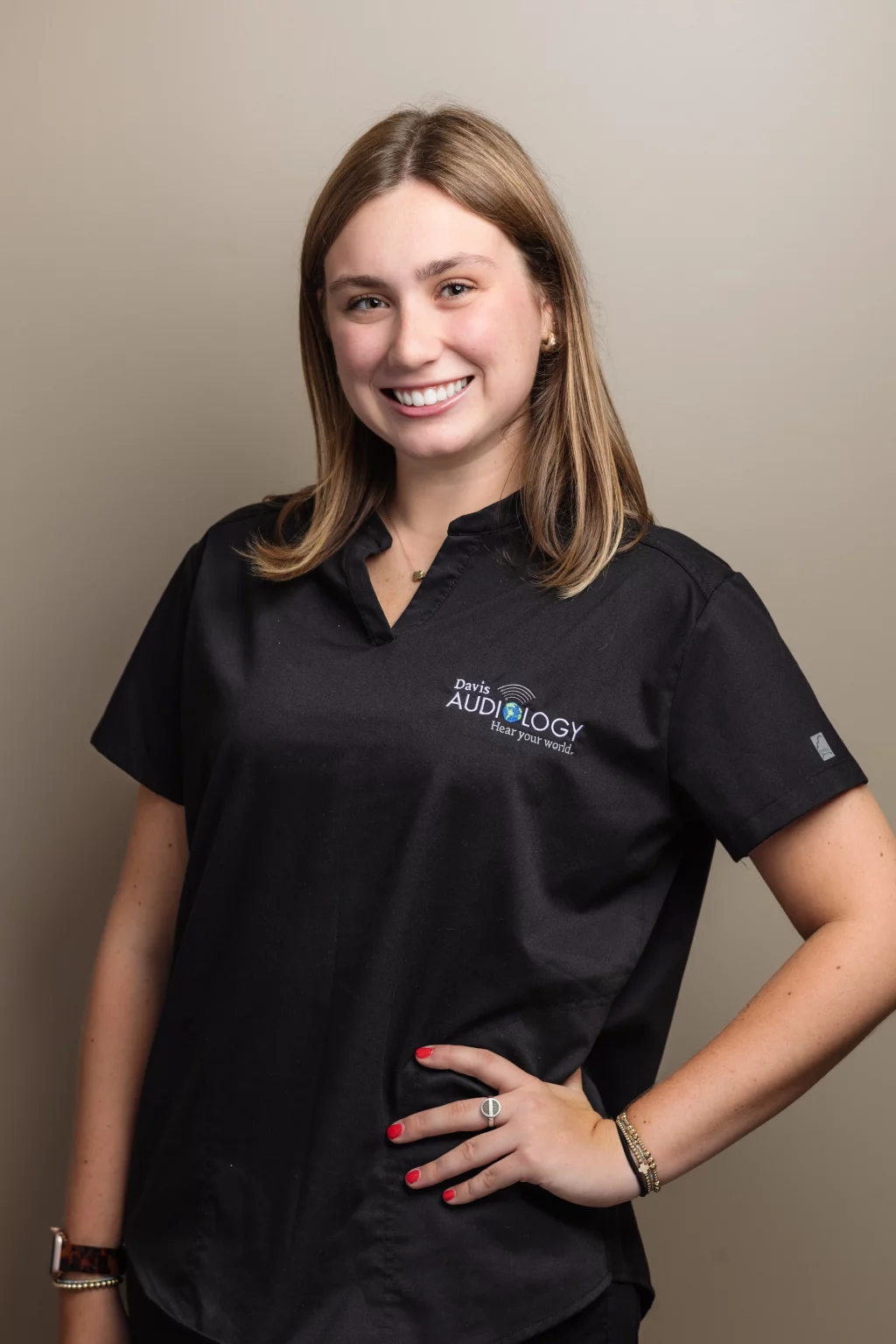 Headshots and branding for Davis Audiology in Taylors SC.: Smiling woman with shoulder-length brown hair wearing a black Davis Audiology scrub top, standing against a neutral background with one hand on her hip.