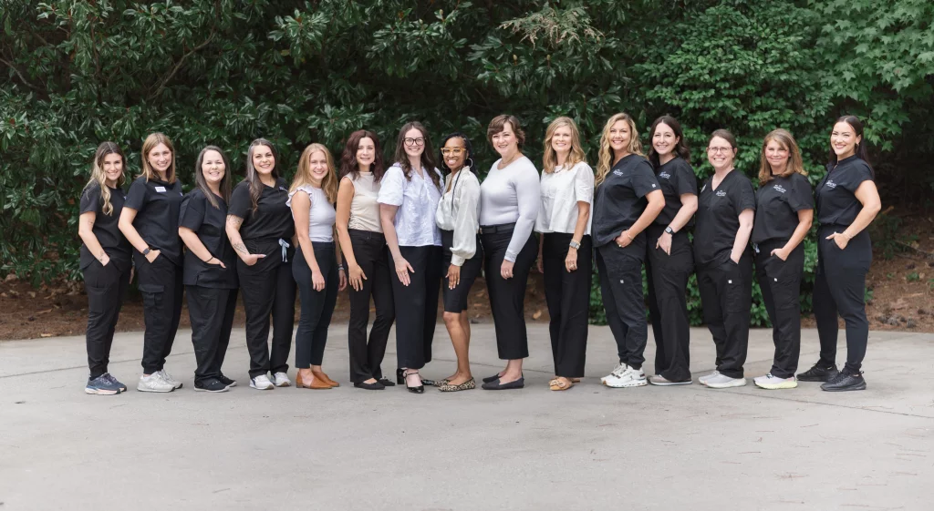Headshots and branding for Davis Audiology in Taylors SC.: Group portrait of 15 women, many wearing black medical scrubs, standing outdoors in front of lush greenery.