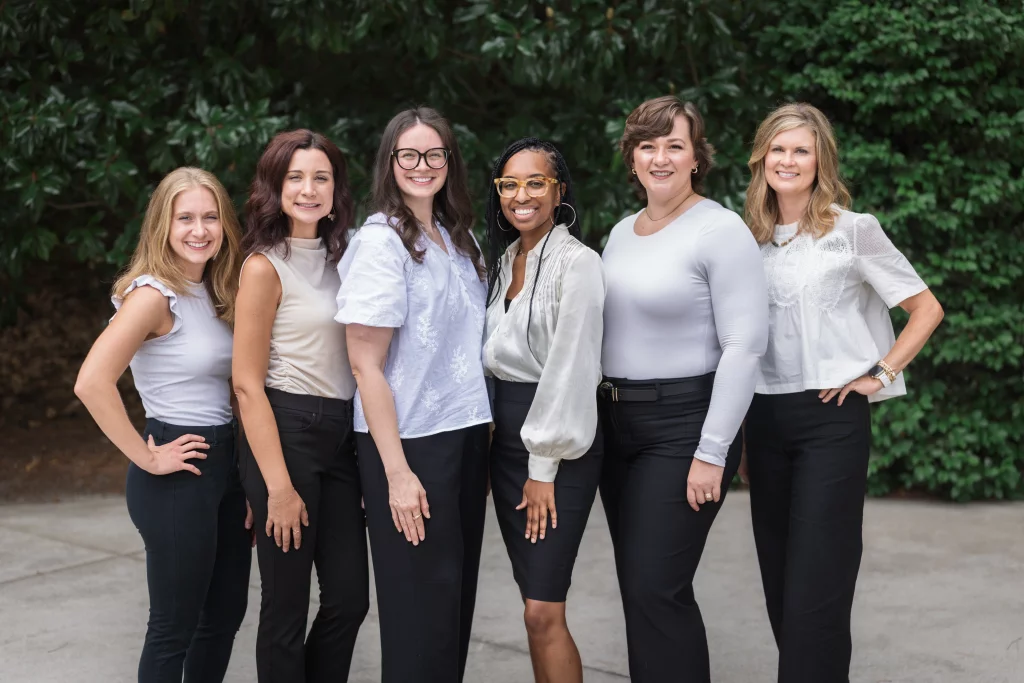Headshots and branding for Davis Audiology in Taylors SC.: Group portrait of six smiling women standing outdoors in front of lush greenery, wearing light-colored tops and dark pants.
