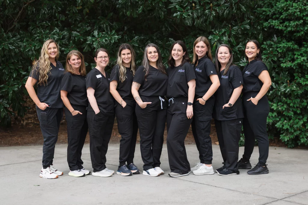 Headshots and branding for Davis Audiology in Taylors SC.: Group portrait of nine women in black medical scrubs standing together outdoors in front of leafy greenery.
