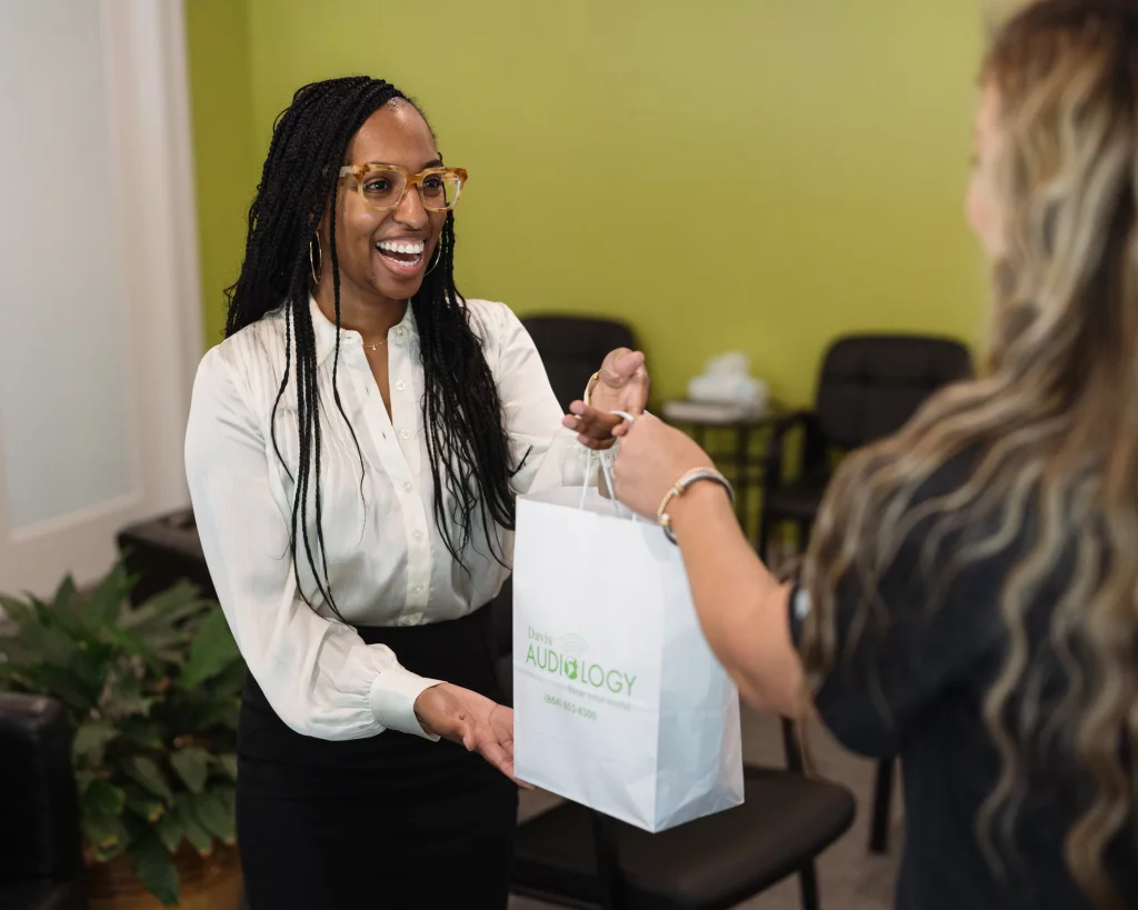 Headshots and branding for Davis Audiology in Taylors SC.: Smiling woman in glasses receives a white Davis Audiology bag from another woman in an office reception area.