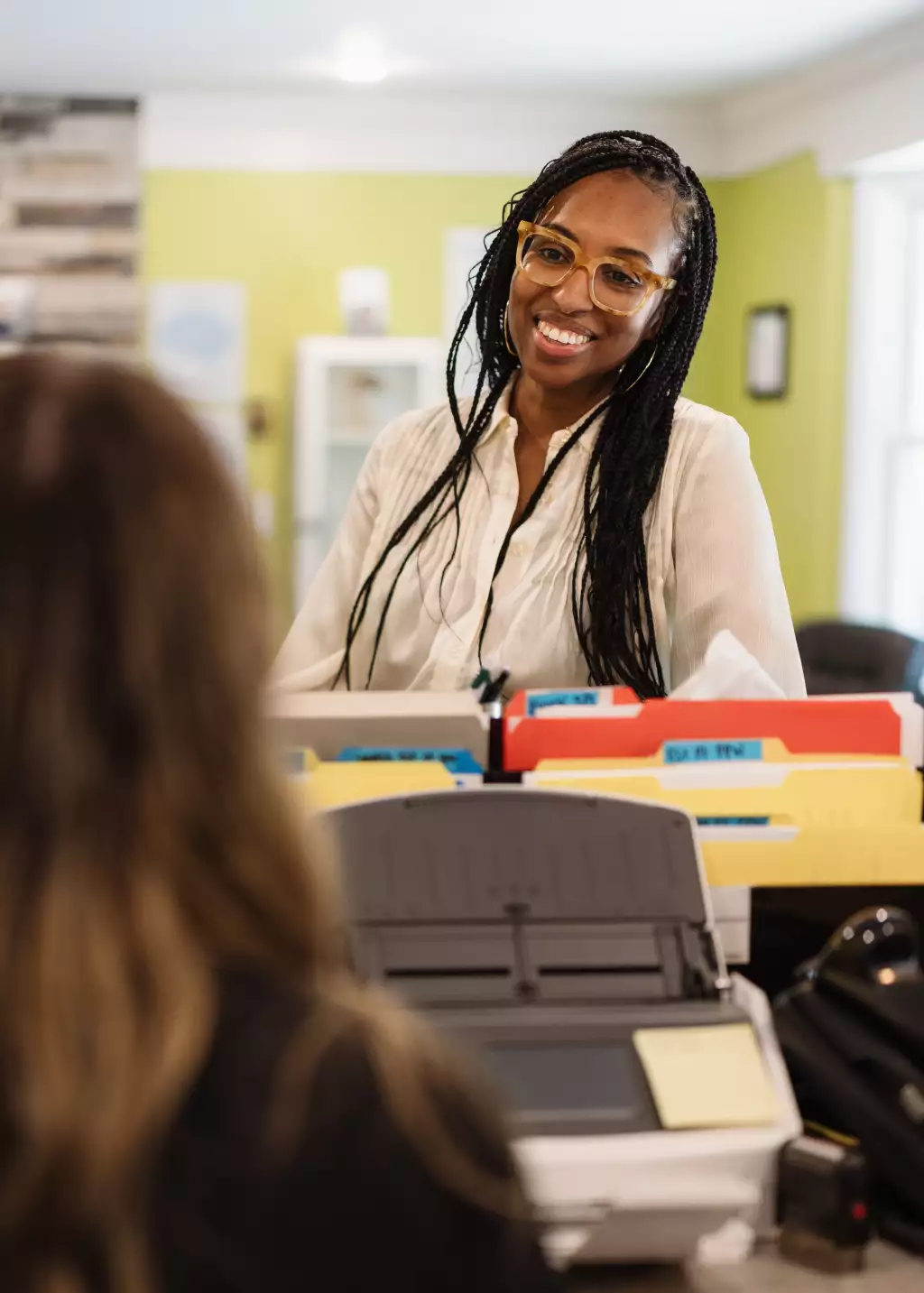 : Smiling woman with long braids and yellow glasses sits behind an office desk, facing a blurred person in the foreground.