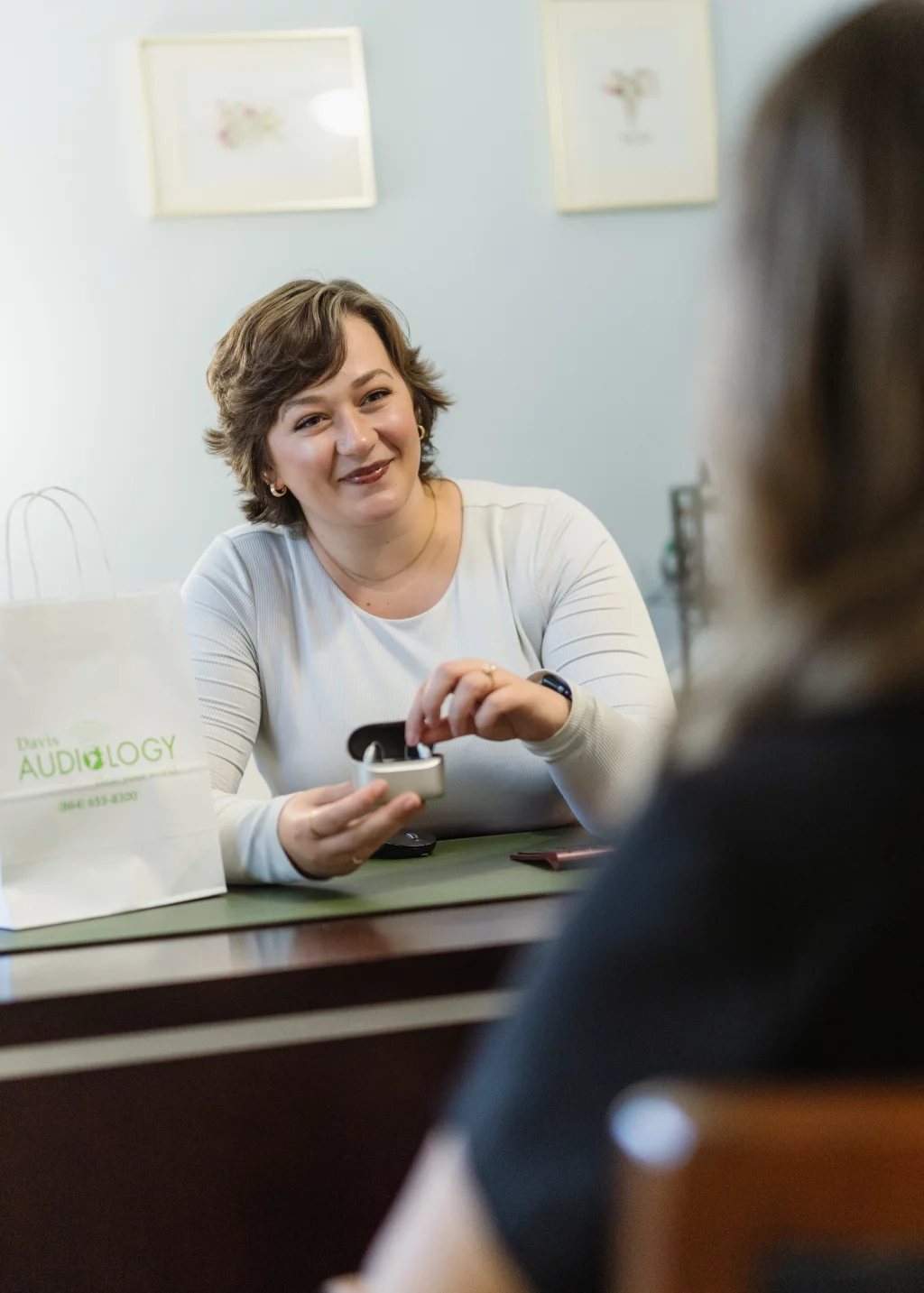 Headshots and branding for Davis Audiology in Taylors SC.: Audiologist smiling across a desk while demonstrating a small hearing device to a client; a bag labeled &ldquo;Paris Audiology&rdquo; sits on the desk.