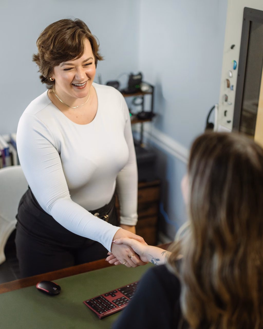 Headshots and branding for Davis Audiology in Taylors SC.: Smiling woman in a white top shakes hands with another person across a desk in an office.