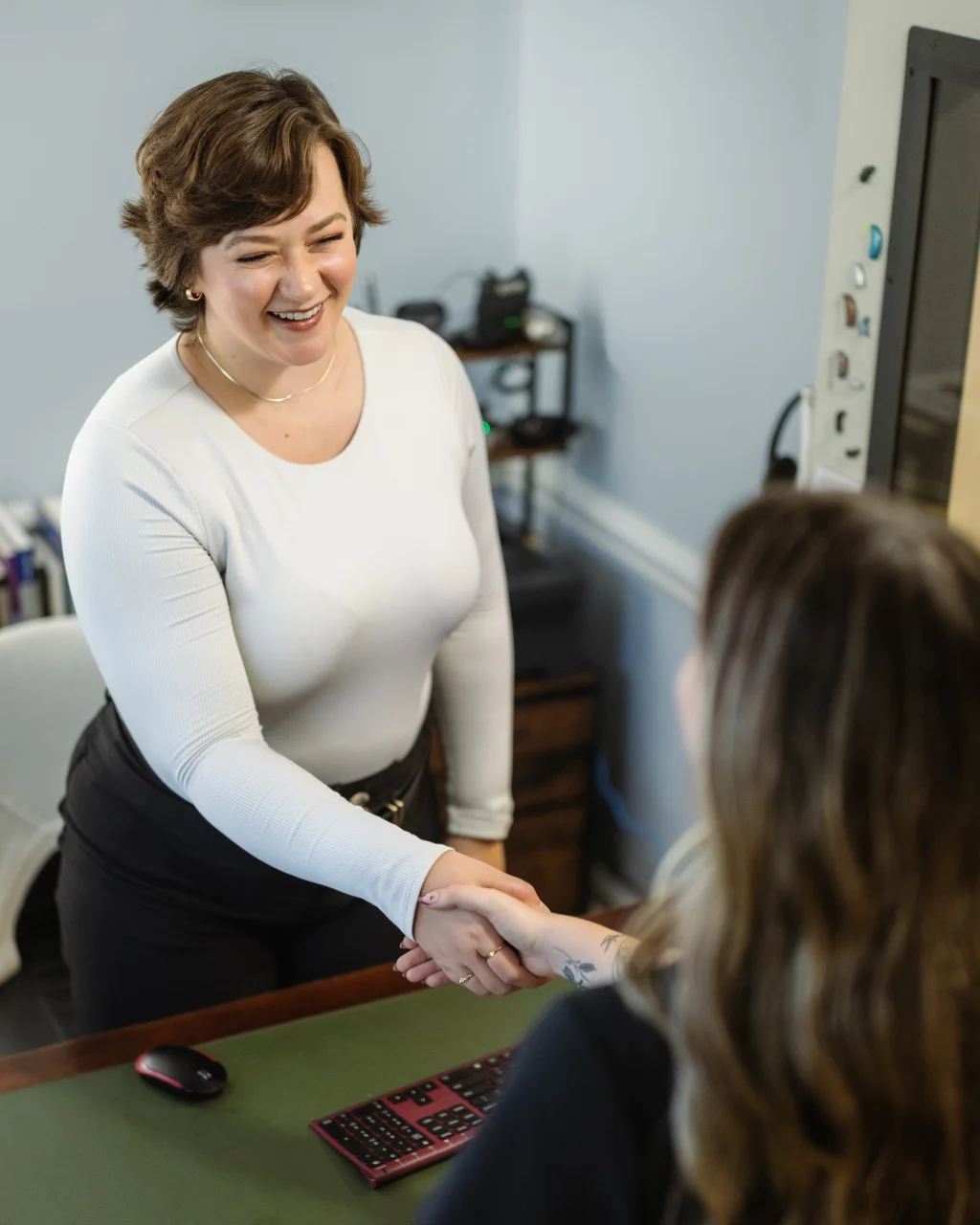Headshots and branding for Davis Audiology in Taylors SC.: Smiling woman in a white top shakes hands with another person across a desk in an office.