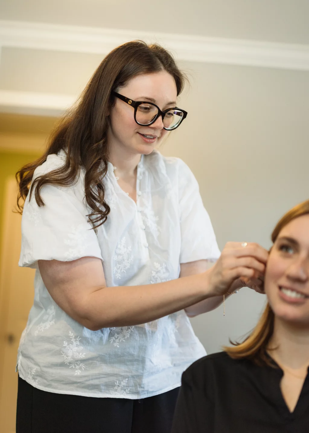 Headshots and branding for Davis Audiology in Taylors SC.: A woman wearing glasses adjusts an earring on a smiling seated woman.