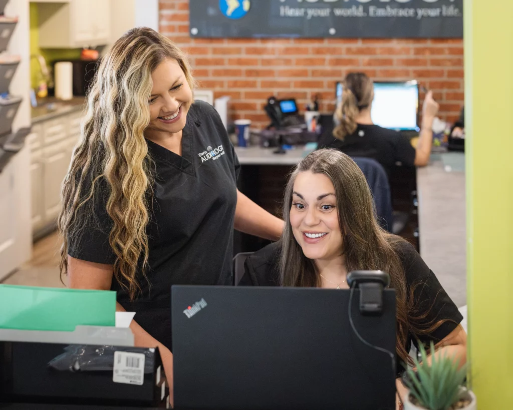Headshots and branding for Davis Audiology in Taylors SC.: Two smiling women work at a reception desk in an audiology office, with computers, equipment, and a brick wall in the background.