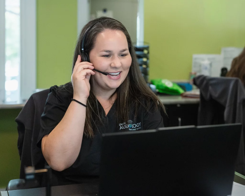 Headshots and branding for Davis Audiology in Taylors SC.: Smiling woman wearing a headset and black &ldquo;Davis Audiology&rdquo; shirt works at a computer in an office.