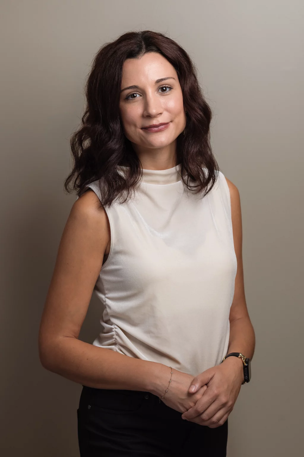 Headshots and branding for Davis Audiology in Taylors SC.: Portrait of a woman with shoulder-length dark wavy hair wearing a white sleeveless top and black pants, standing against a neutral background with her hands clasped.