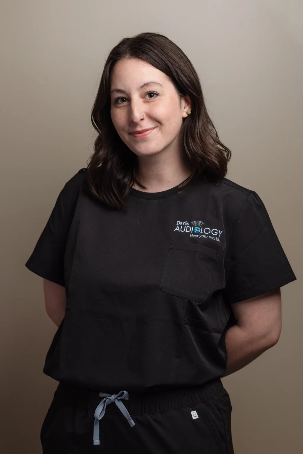 Headshots and branding for Davis Audiology in Taylors SC.: Portrait of a smiling woman with shoulder-length dark hair wearing black medical scrubs embroidered with the Davis Audiology logo.