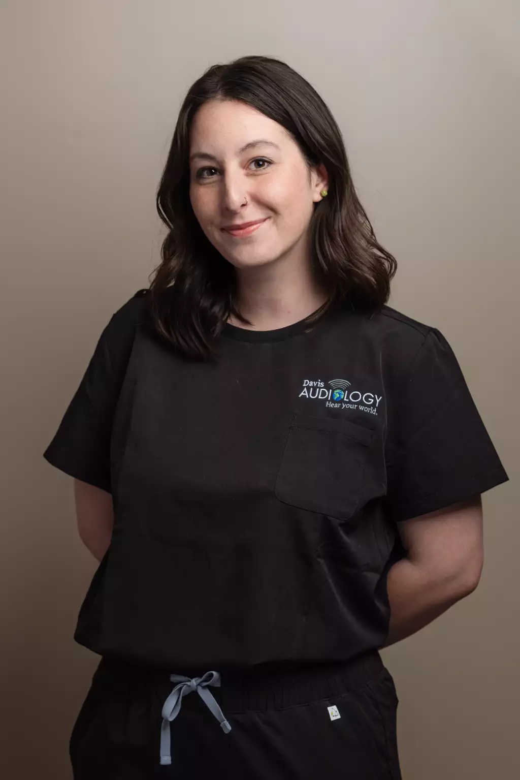 : Portrait of a smiling woman with shoulder-length dark hair wearing black medical scrubs embroidered with the Davis Audiology logo.