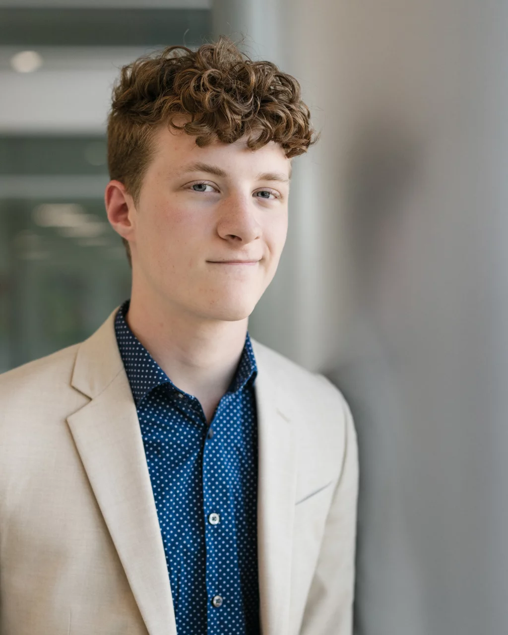 Jacobi's senior photos taken at Falls Park, Greenville SC: Young man with curly brown hair wearing a beige blazer and blue polka dot shirt, standing indoors and looking at the camera with a slight smile.