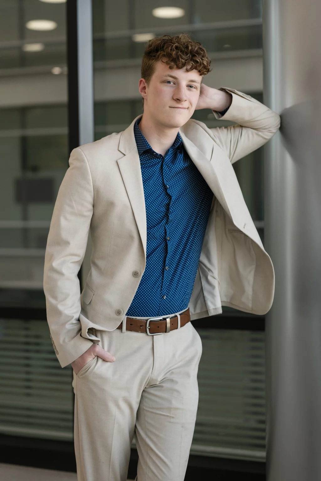 Jacobi's senior photos taken at Falls Park, Greenville SC: Young man with curly hair wearing a light beige suit and blue patterned shirt, standing indoors with one hand in his pocket and the other resting behind his head against a wall.