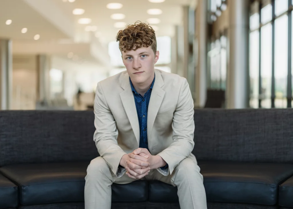 Jacobi's senior photos taken at Falls Park, Greenville SC: Young man with curly hair wearing a light beige suit and blue shirt, sitting on a dark sofa in a modern, well-lit interior with large windows.