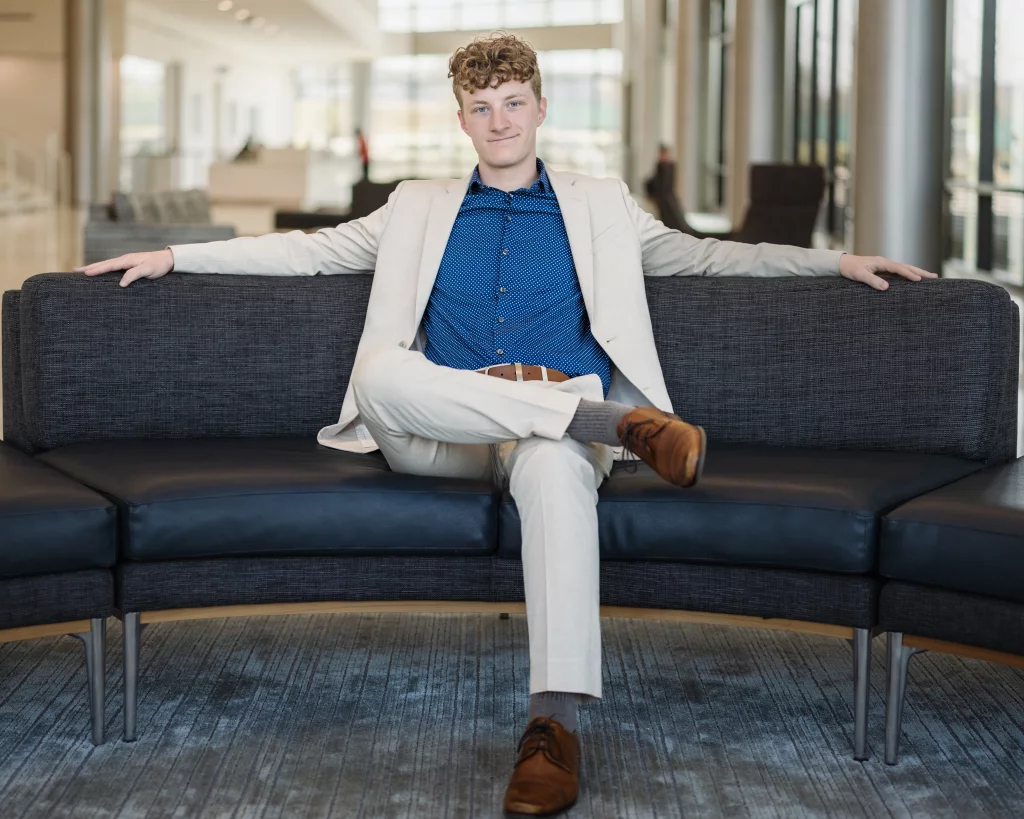 Jacobi's senior photos taken at Falls Park, Greenville SC: Young man with curly hair in a light suit and blue shirt sits confidently on a modern, curved black sofa in a bright, spacious interior.