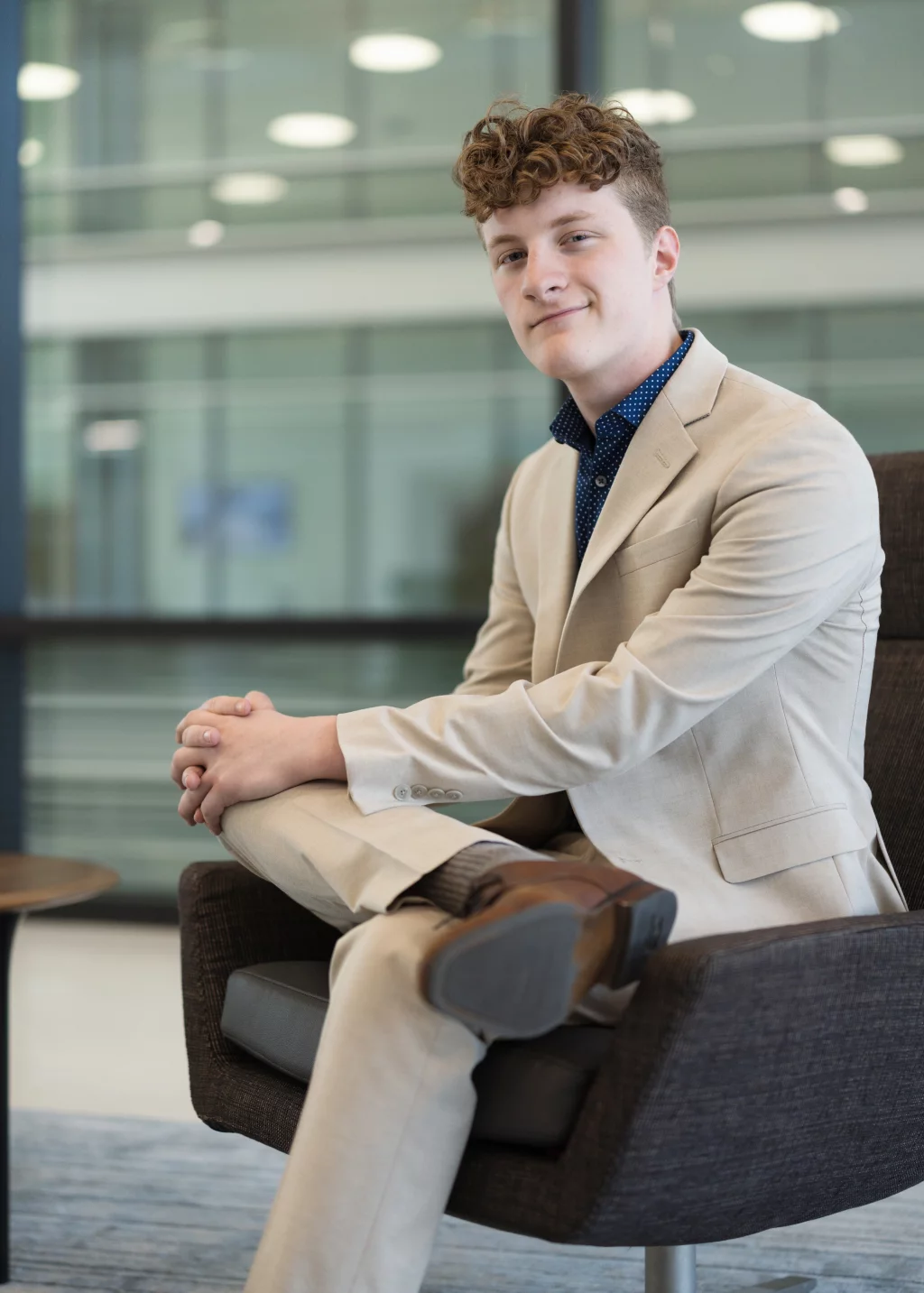 Jacobi's senior photos taken at Falls Park, Greenville SC: Young man with curly hair wearing a beige suit and brown shoes, sitting cross-legged in a modern chair, smiling slightly in a bright, contemporary indoor setting.