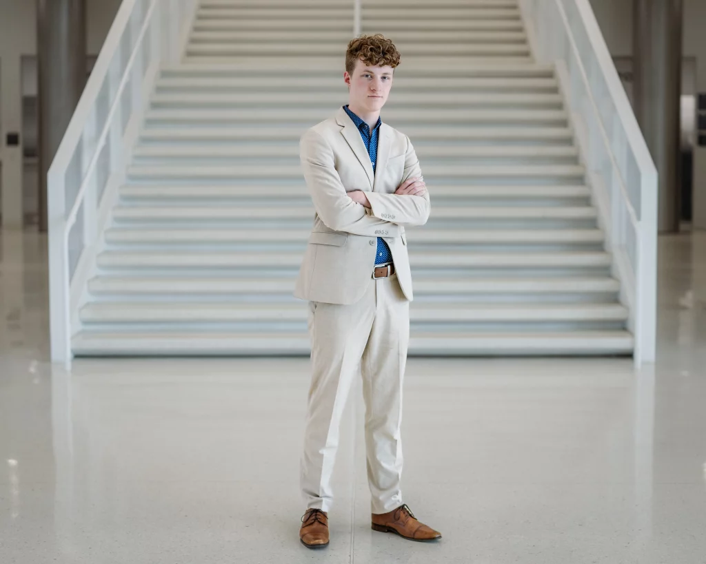 Jacobi's senior photos taken at Falls Park, Greenville SC: A young man in a light-colored suit stands with arms crossed in front of a wide staircase in a modern, bright interior space.