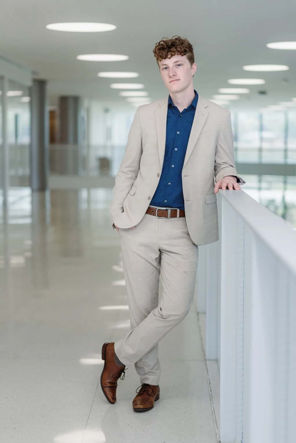 Jacobi's senior photos taken at Falls Park, Greenville SC: Young man in a light beige suit and blue patterned shirt leaning casually against a white railing in a bright, modern hallway with circular ceiling lights.