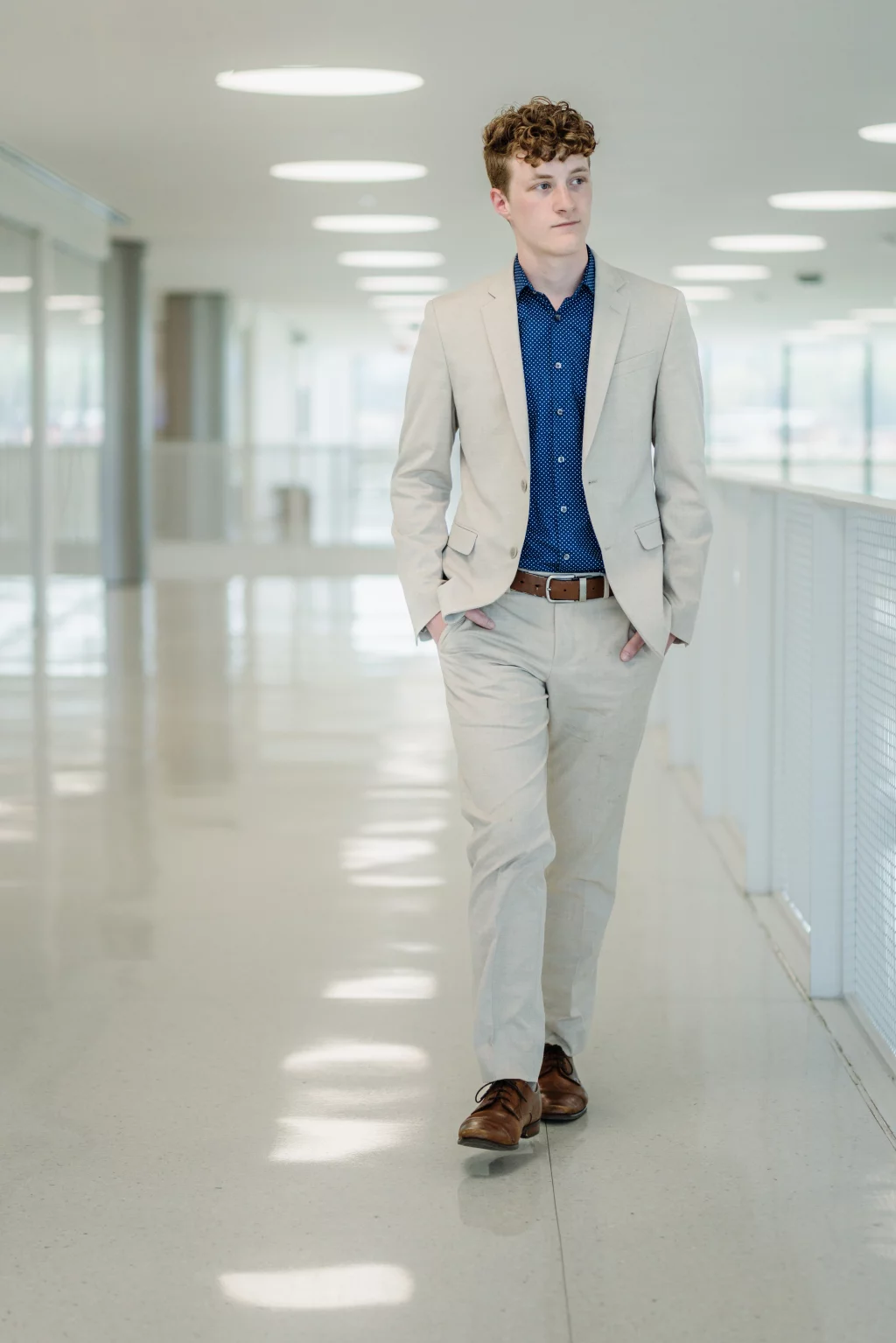 Jacobi's senior photos taken at Falls Park, Greenville SC: Young man with curly hair wearing a light beige suit, blue patterned shirt, and brown shoes walks confidently down a bright, modern hallway with hands in pockets.