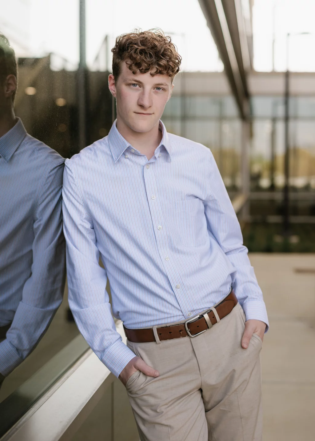 Jacobi's senior photos taken at Falls Park, Greenville SC: Young person with curly hair wearing a light blue striped button-up shirt and beige pants, standing with hands in pockets and leaning against a reflective glass wall outdoors.