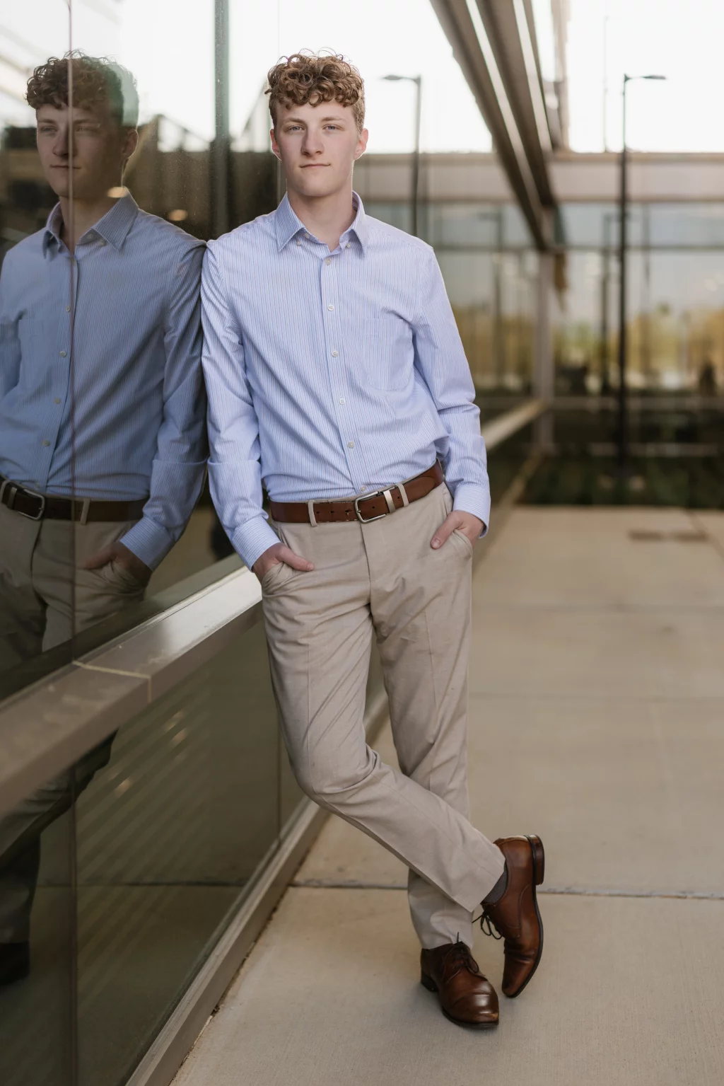 Jacobi's senior photos taken at Falls Park, Greenville SC: Young man with curly hair wearing a light blue dress shirt, tan pants, and brown shoes stands casually with hands in pockets, leaning against a glass wall with his reflection visible.