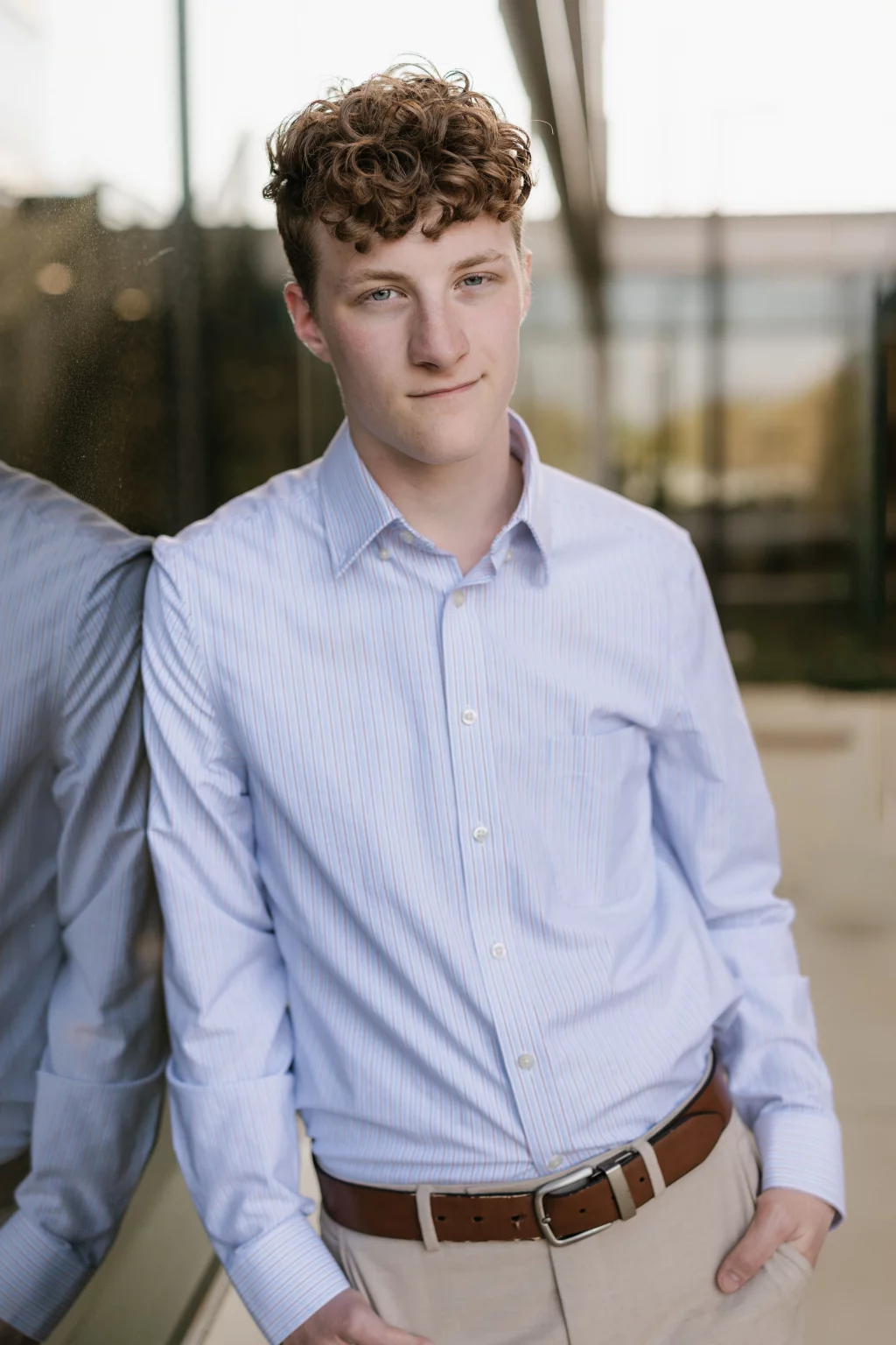 Jacobi's senior photos taken at Falls Park, Greenville SC: Teen boy with curly hair wearing a light blue striped dress shirt and tan pants, standing outdoors with his back against a reflective glass wall, looking at the camera with a slight smile.