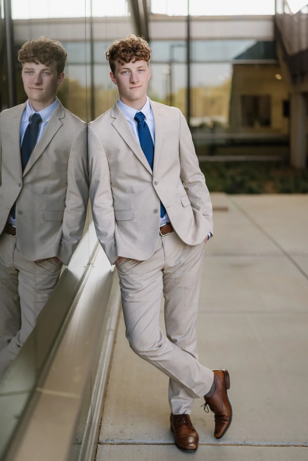 Jacobi's senior photos taken at Falls Park, Greenville SC: Young man in a light beige suit with a blue tie and brown dress shoes, standing with hands in pockets and leaning against a glass wall outside a modern building, with his reflection visible in the glass.