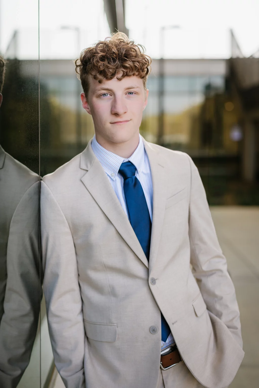 Jacobi's senior photos taken at Falls Park, Greenville SC: Young man with curly hair wearing a light beige suit, blue tie, and striped shirt, standing outdoors and leaning against a reflective glass wall.