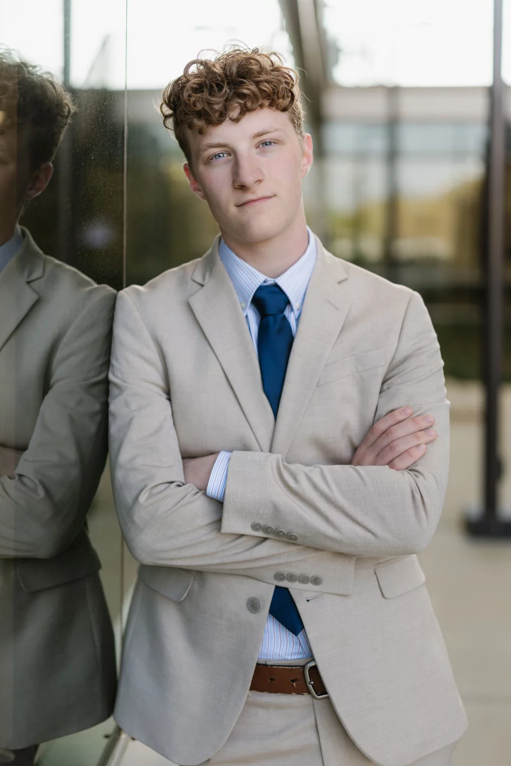 Jacobi's senior photos taken at Falls Park, Greenville SC: Young person with curly hair wearing a light beige suit, blue tie, and striped shirt, standing with arms crossed and leaning against a reflective glass wall.