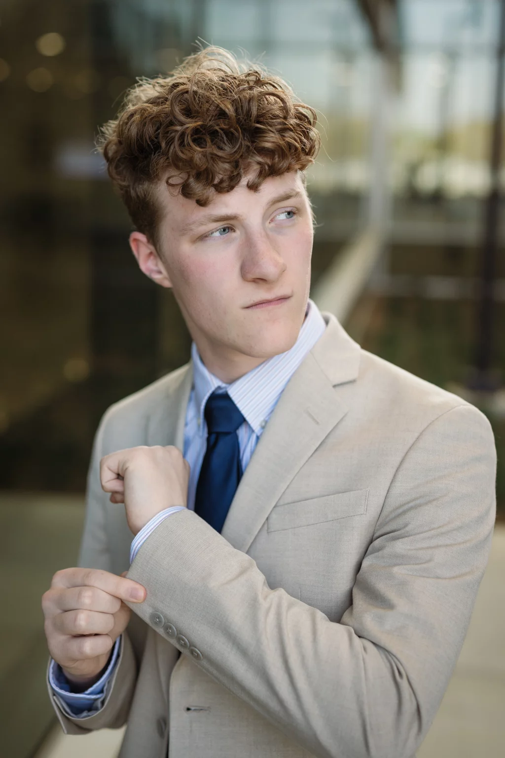 Jacobi's senior photos taken at Falls Park, Greenville SC: Young man with curly hair in a tan suit and blue tie adjusting his sleeve, looking to the side with a serious expression.