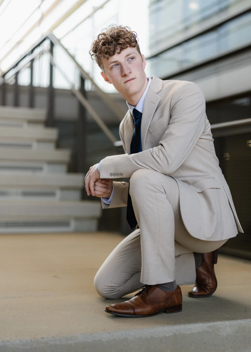 Jacobi's senior photos taken at Falls Park, Greenville SC: Young man with curly hair wearing a light beige suit, blue tie, and brown dress shoes, kneeling on one knee near a modern staircase with glass and metal railing.