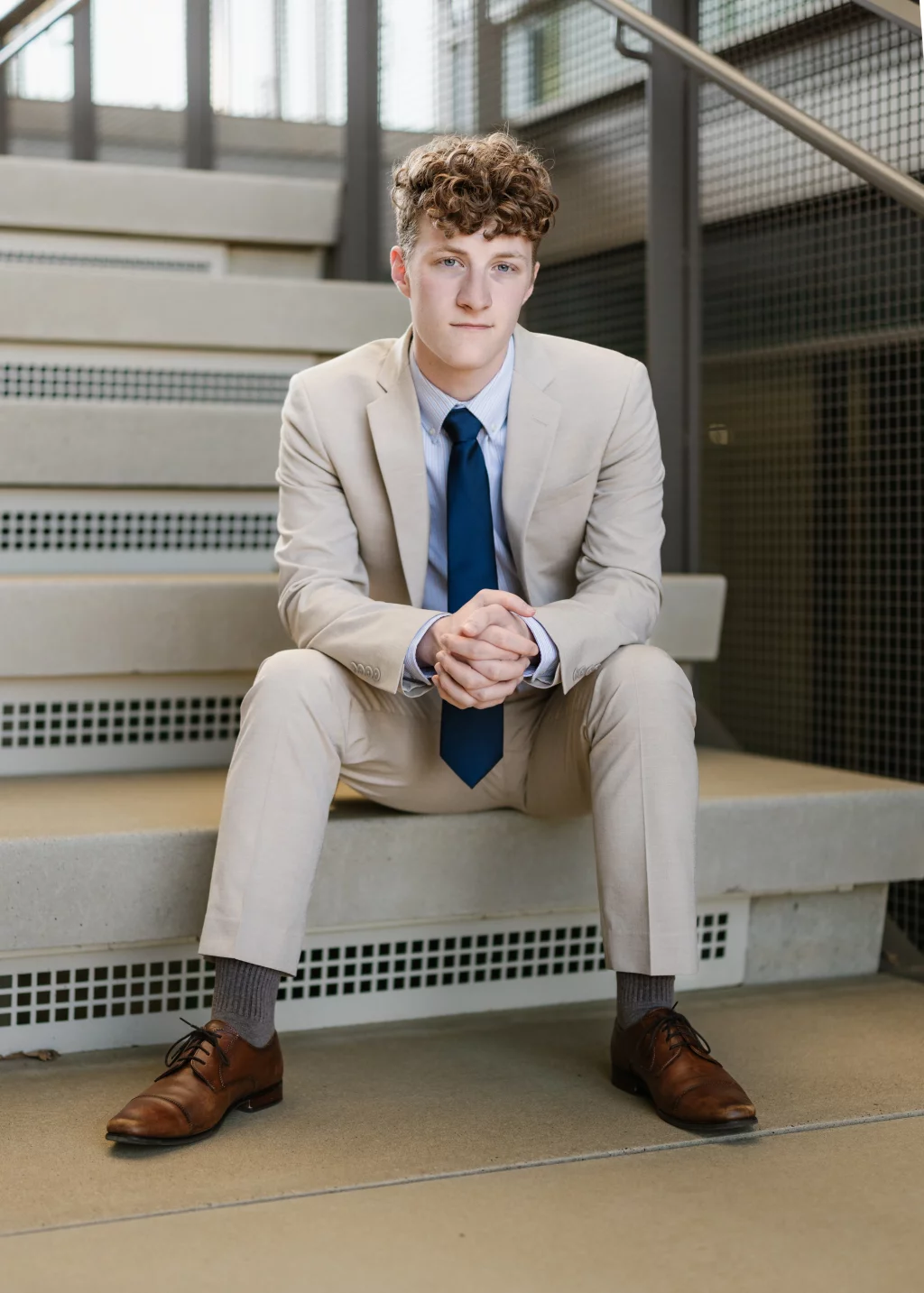 Jacobi's senior photos taken at Falls Park, Greenville SC: Young man with curly hair wearing a light beige suit, blue tie, and brown dress shoes, sitting on concrete steps indoors with hands clasped.