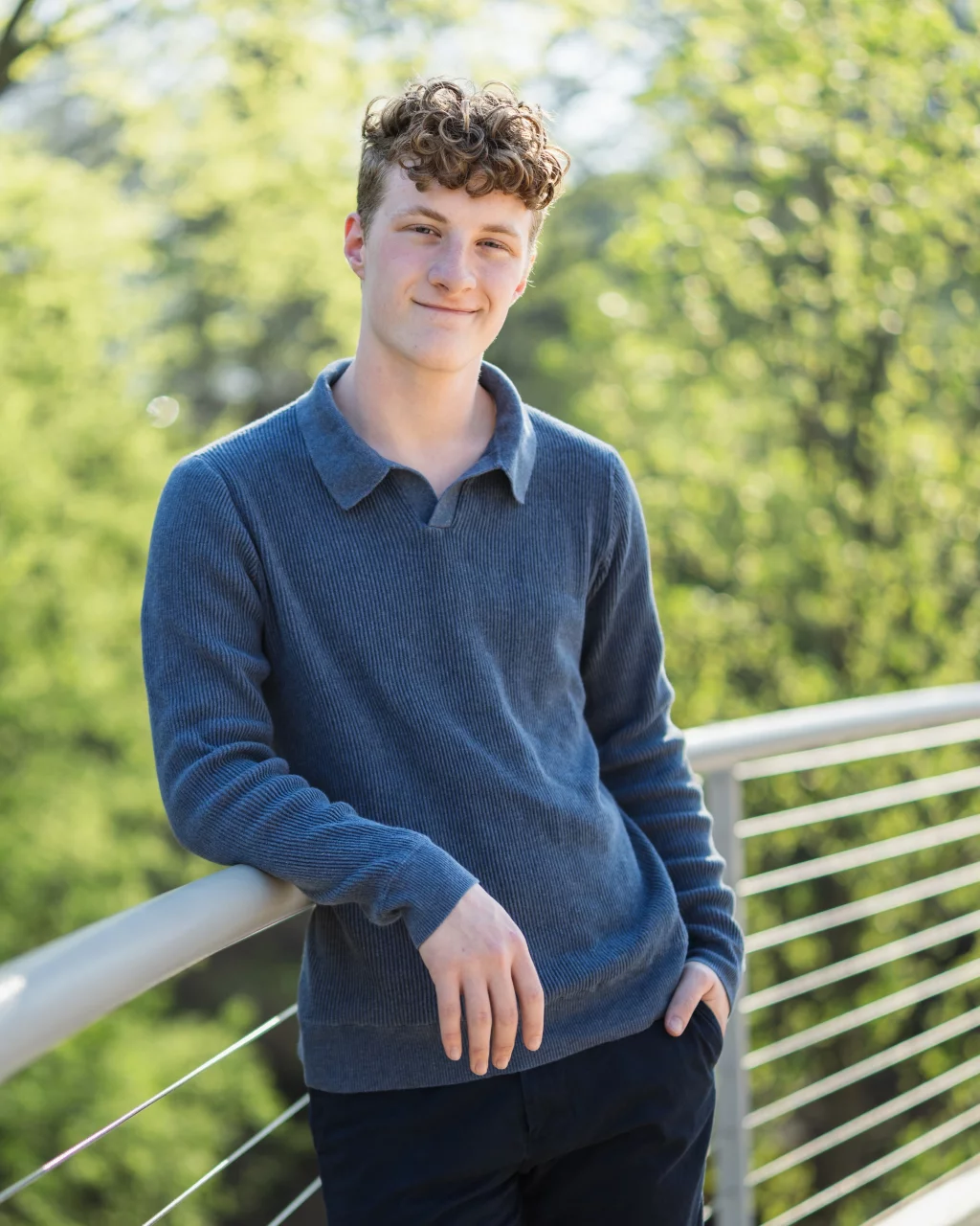 Jacobi's senior photos taken at Falls Park, Greenville SC: Young man with curly hair wearing a blue sweater, leaning on a metal railing outdoors with green trees in the background.