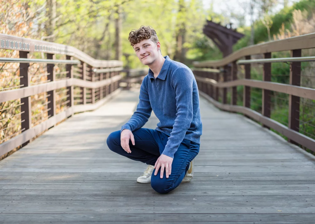 Jacobi's senior photos taken at Falls Park, Greenville SC: A young man with curly hair, wearing a blue sweater and pants, kneels and smiles on a wooden bridge surrounded by greenery.