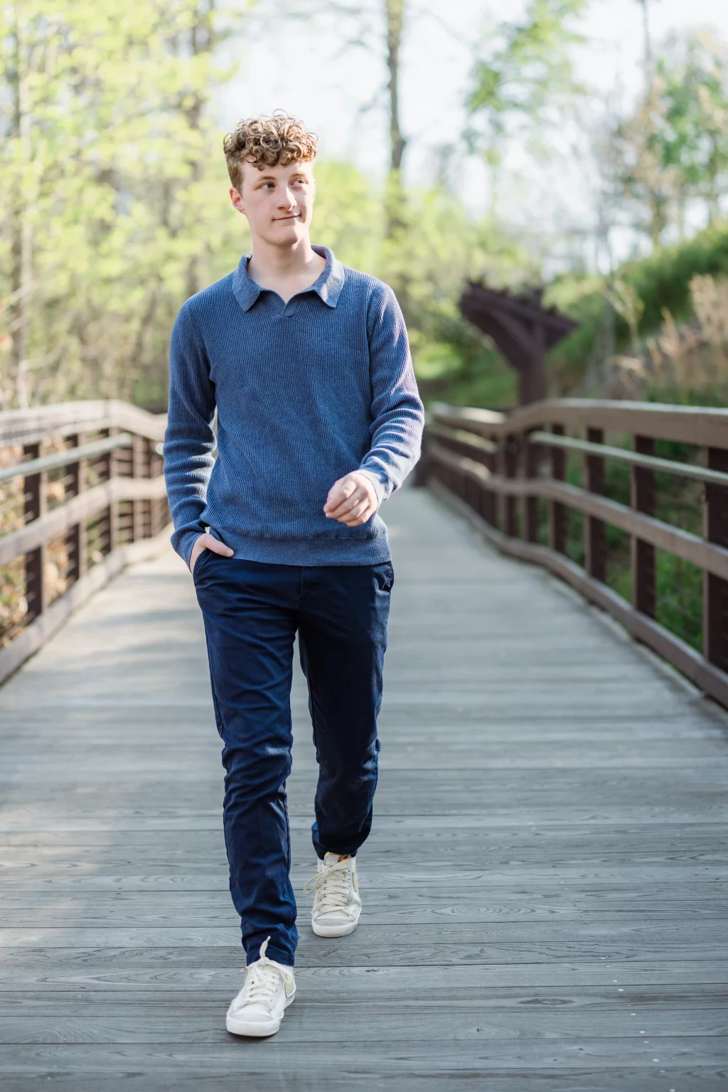 Jacobi's senior photos taken at Falls Park, Greenville SC: Young man with curly hair wearing a blue sweater, navy pants, and white sneakers walking on a wooden bridge surrounded by trees.