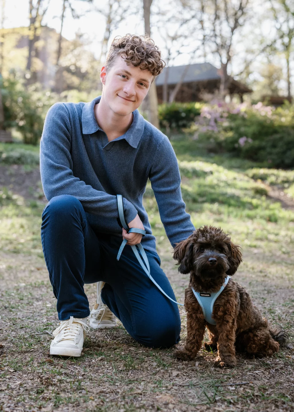 Jacobi's senior photos taken at Falls Park, Greenville SC: A young person kneels outdoors on one knee, holding a leash and smiling at the camera, next to a small brown dog wearing a blue harness. Trees and greenery are in the background.